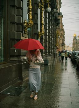 A woman walks on a rainy sidewalk, sheltered by a vibrant red umbrella, in Saint Petersburg, Russia.