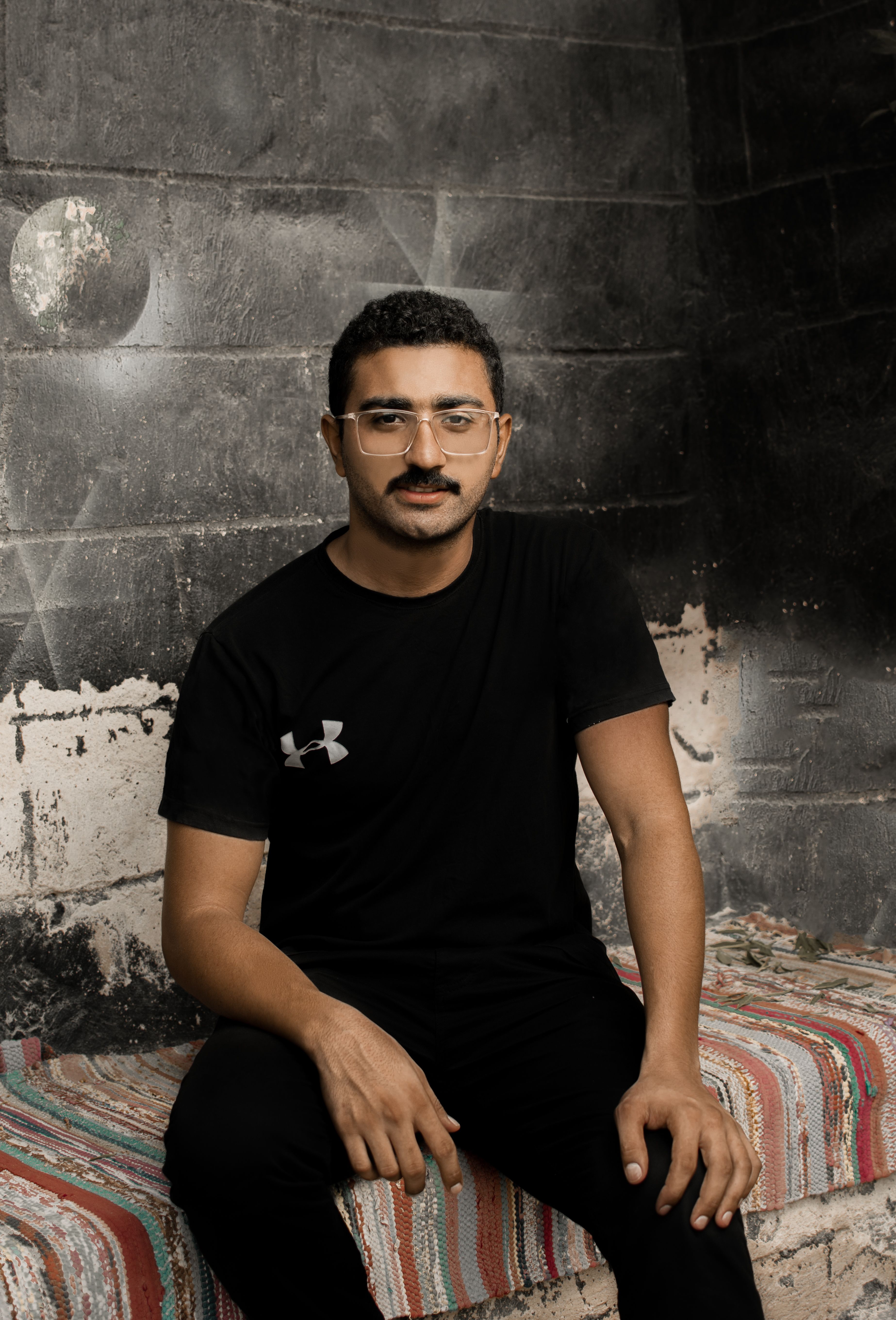 Portrait of a man sitting on a colorful cloth against a grunge wall, wearing black attire.