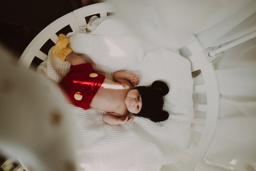 Cute baby wearing mouse ears hat and knitted shorts, lying in a white crib.