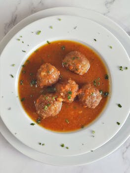 Top-view of homemade meatballs in savory soup garnished with herbs on a white plate.