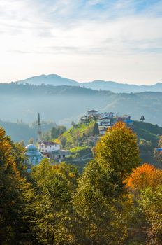 Idyllic mountain town nestled in a vibrant autumn forest with distant peaks under a clear sky.