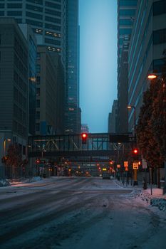 Quiet city street at dusk with snow, high-rise buildings and illuminated traffic lights.