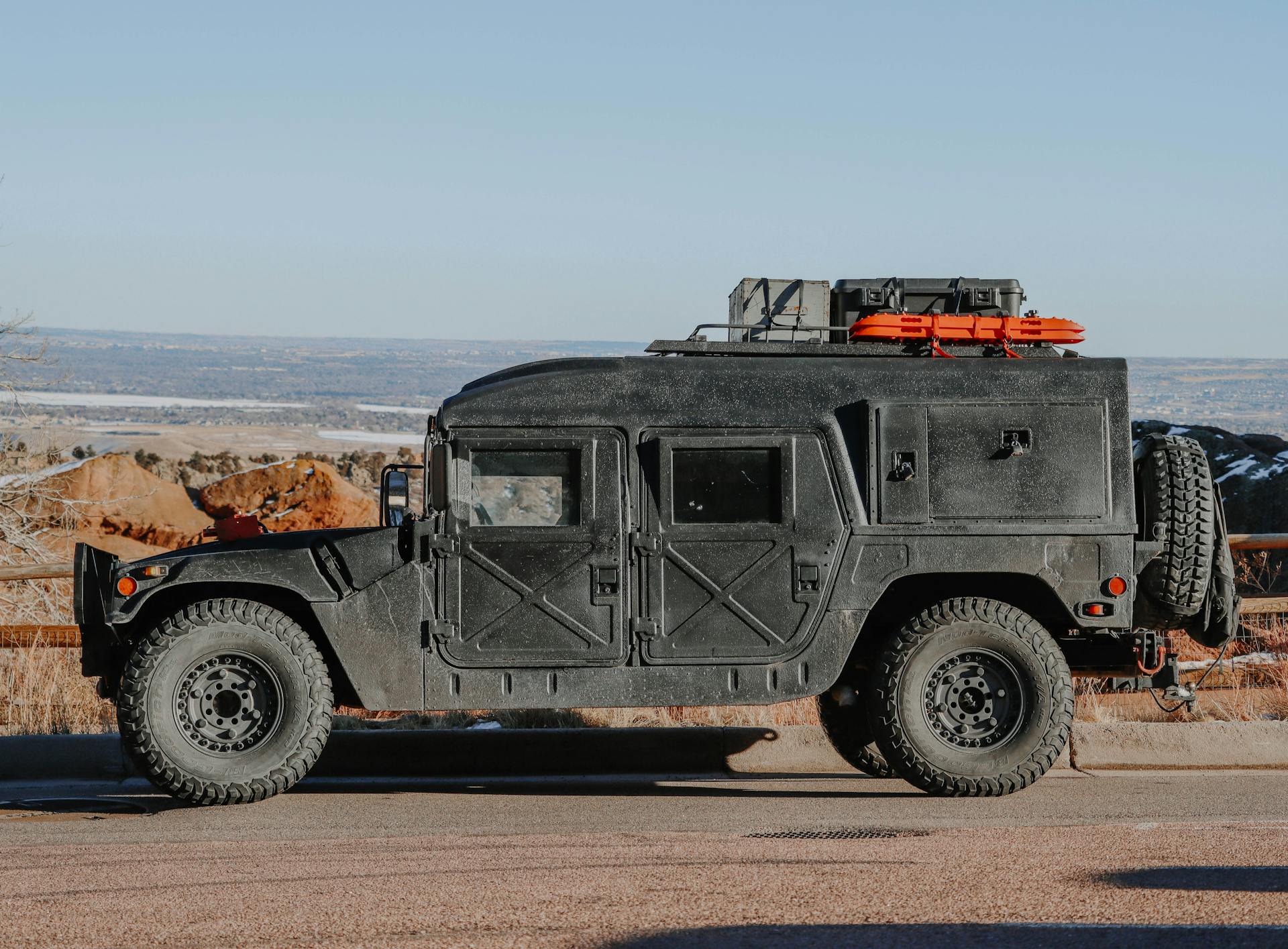 A black armored Humvee with rooftop cargo parked by a scenic mountain road.