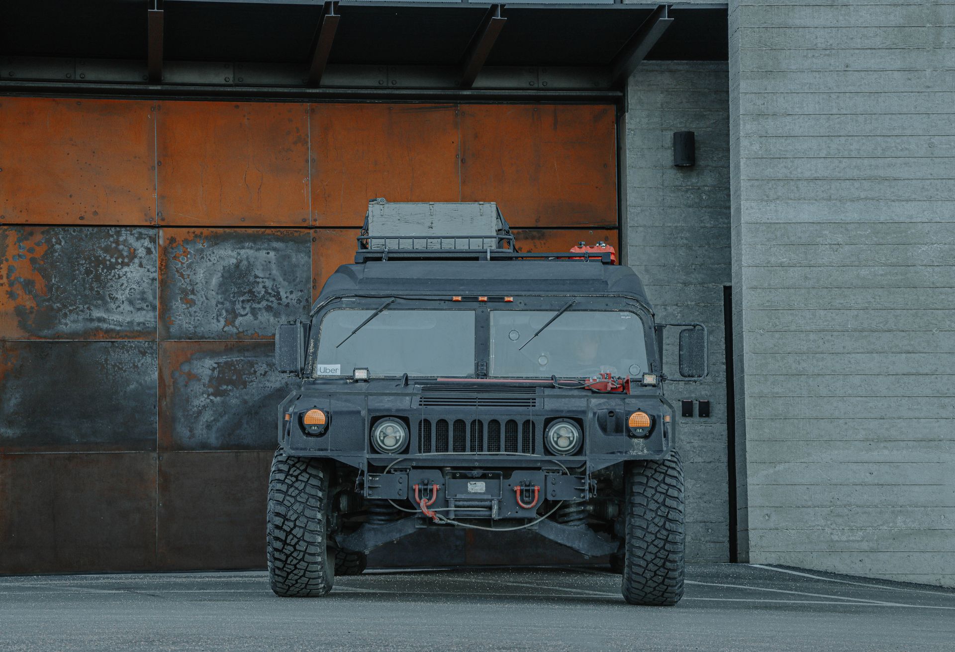 Front view of a rugged black military vehicle parked in an industrial environment with concrete and rust textures.