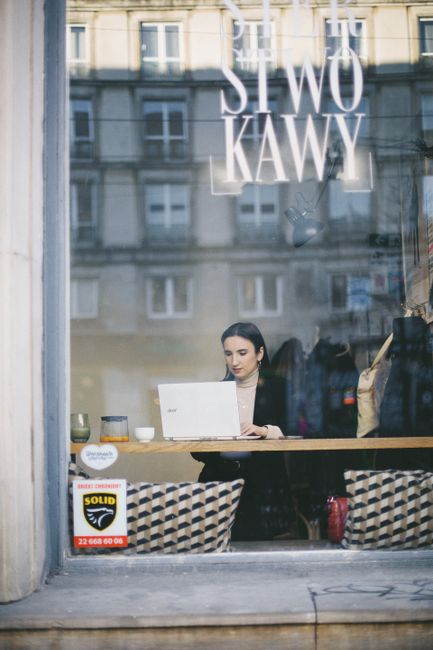 A woman working on a laptop inside a Warsaw café, viewed through a window.