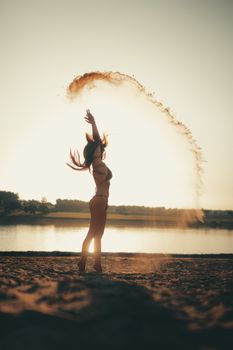 A woman in swimsuit tossing sand playfully on a serene beach at sunset.