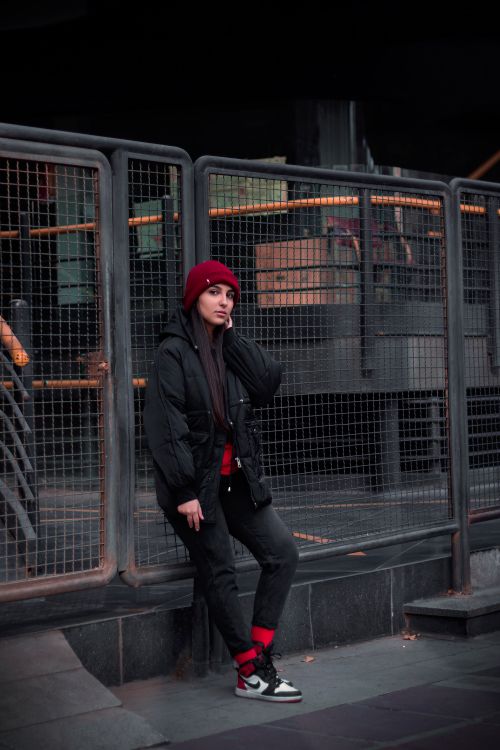 Free Stylish woman leans against urban mesh fence in Tehran, wearing red beanie and black jacket. Stock Photo