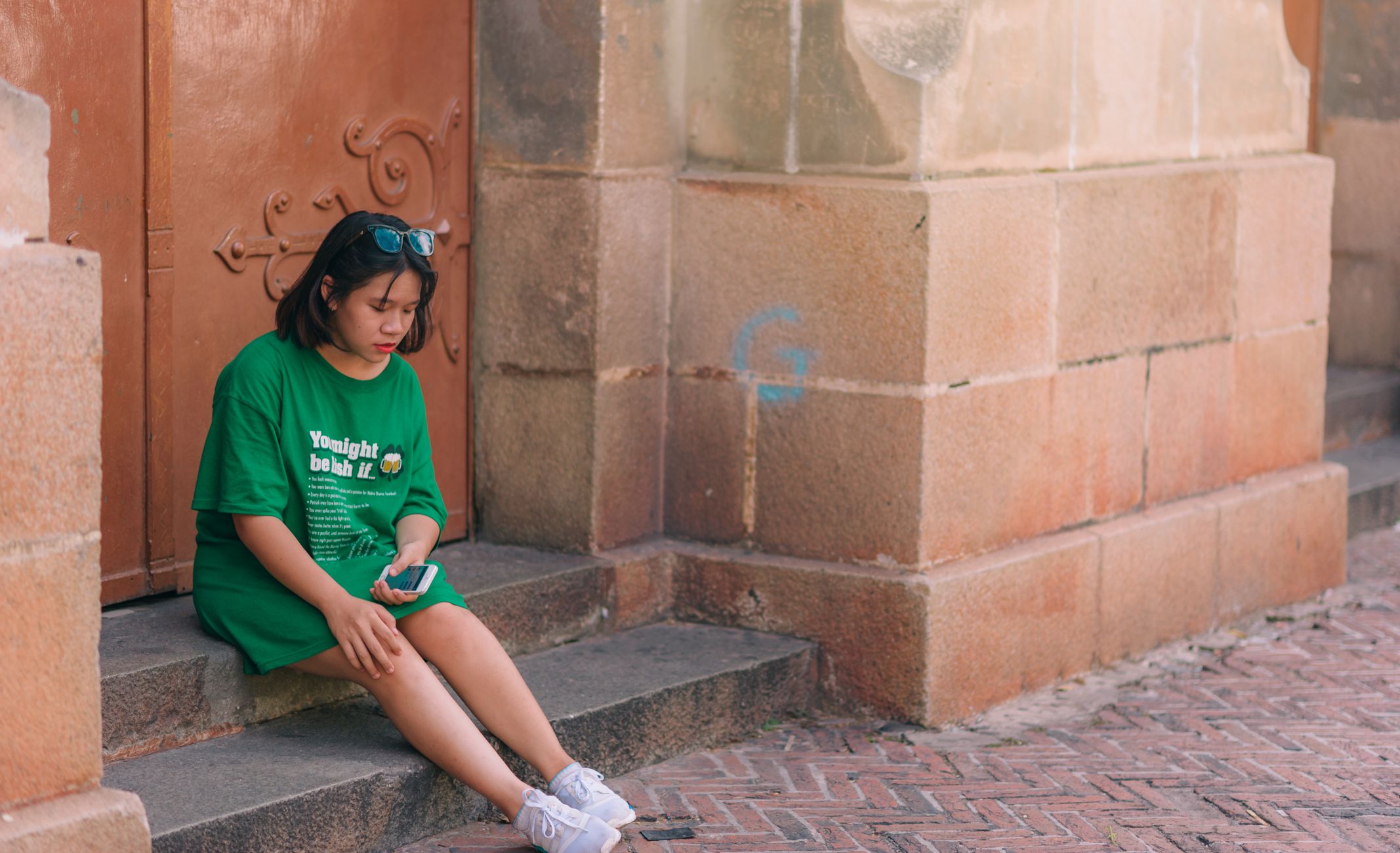 A young woman in a green shirt sits on steps, looking at her phone.