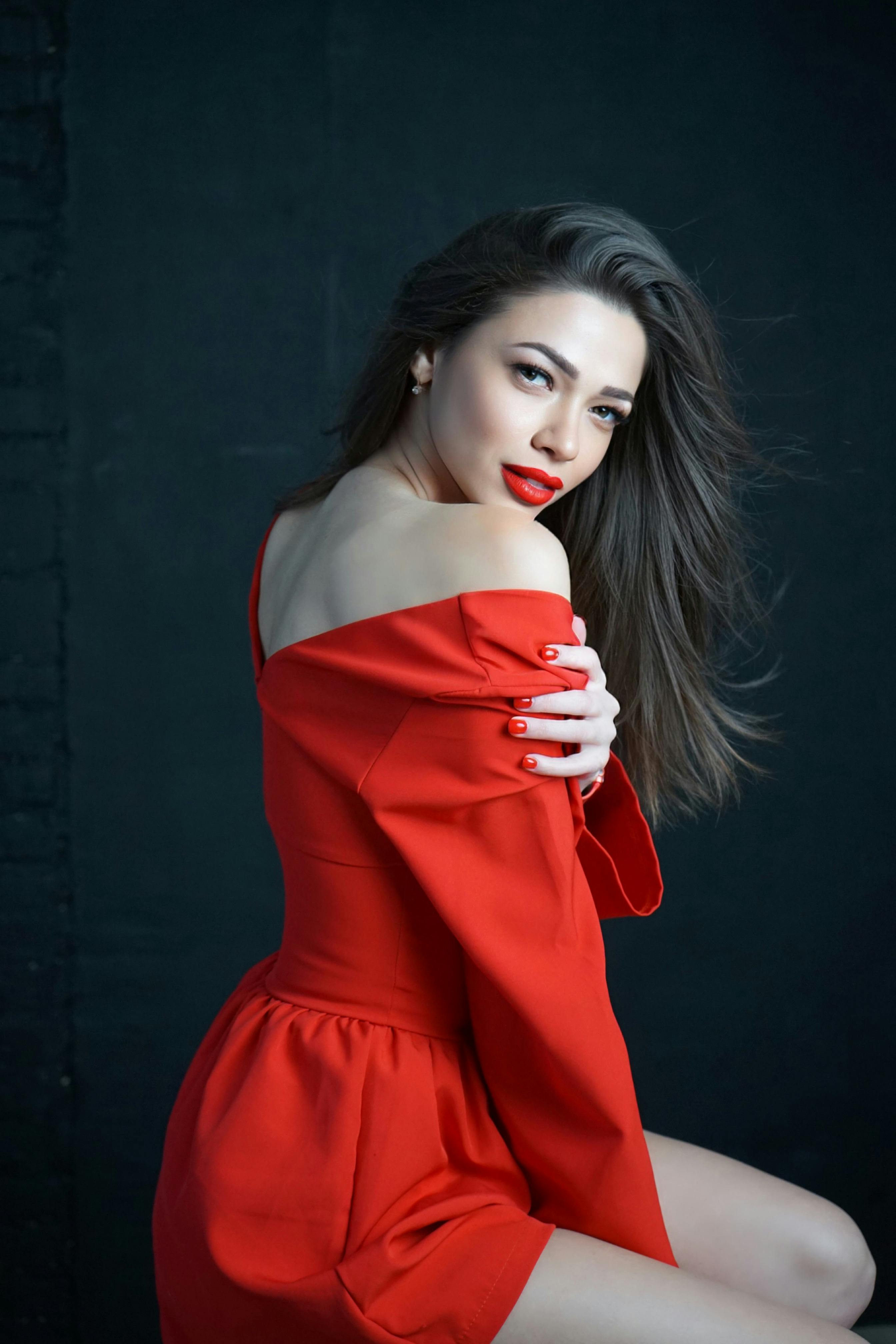 Portrait of a woman with long brown hair in a red dress and lipstick, posing elegantly in a studio.