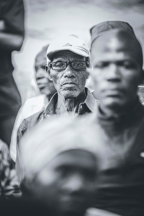 Free A black and white close-up portrait of an elderly man wearing glasses and a cap, surrounded by others. Stock Photo