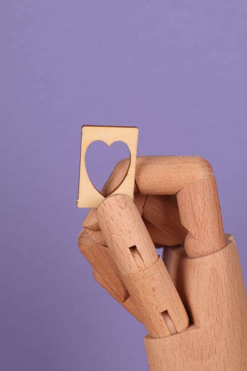 Free A wooden mannequin hand holding a heart-shaped cutout against a purple background, symbolizing love and creativity. Stock Photo