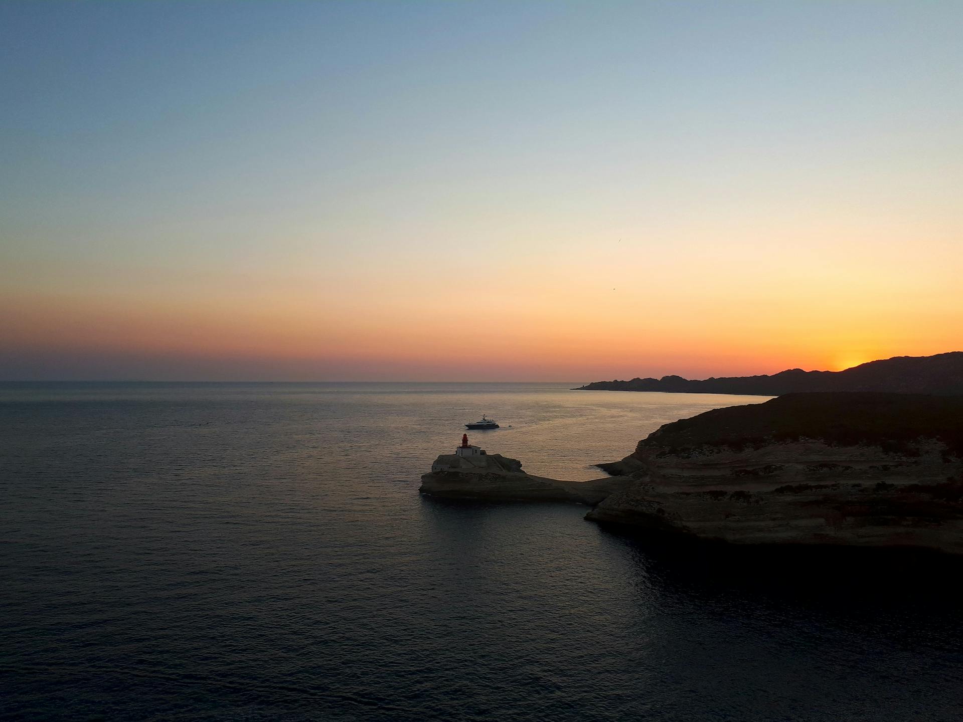 A scenic sunset view of a lighthouse on the Greek coastline with a boat in the horizon.