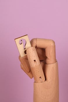 A wooden hand model holding a question mark cutout against a pink background.