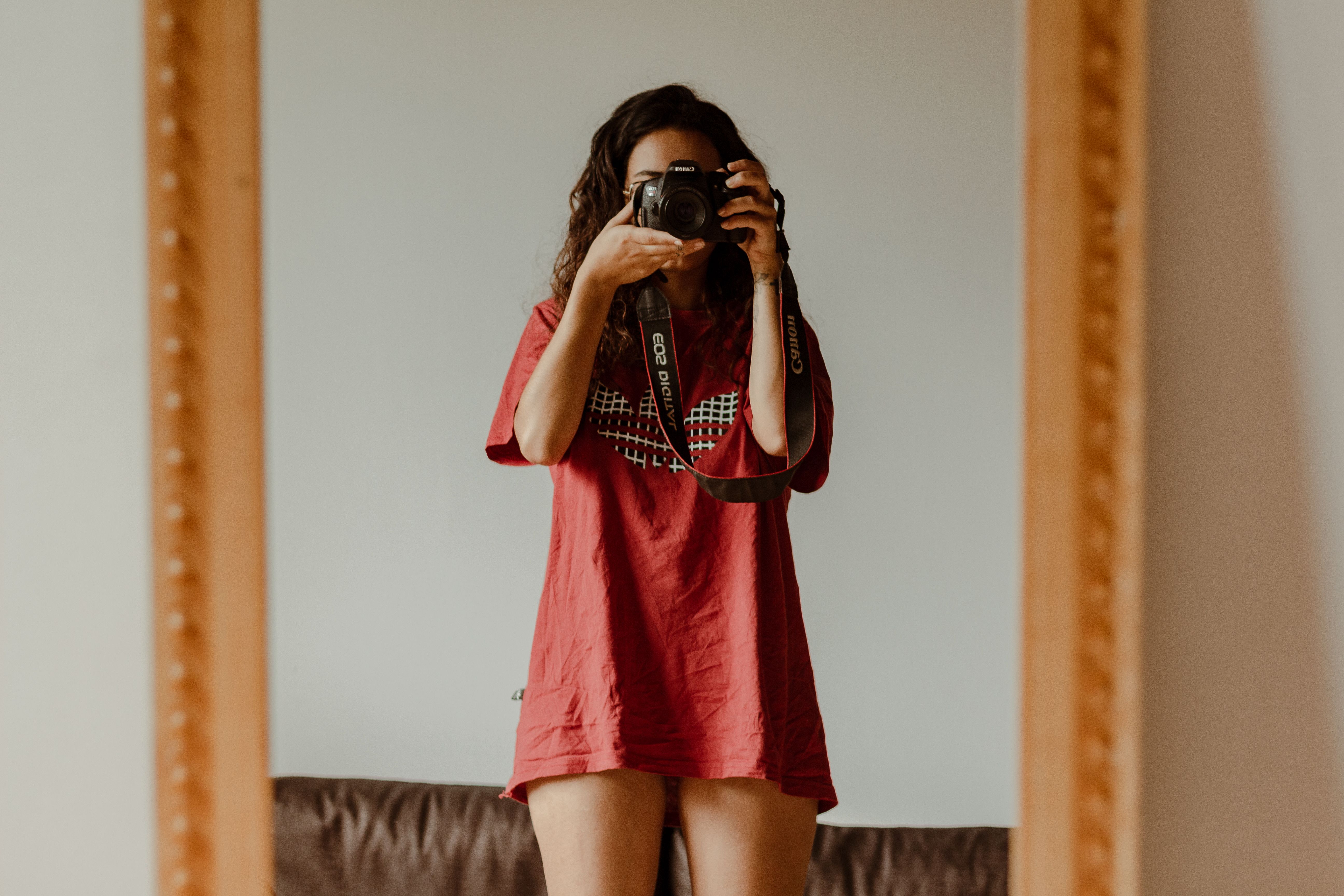 A woman in a red t-shirt taking a selfie in the mirror with a DSLR camera.