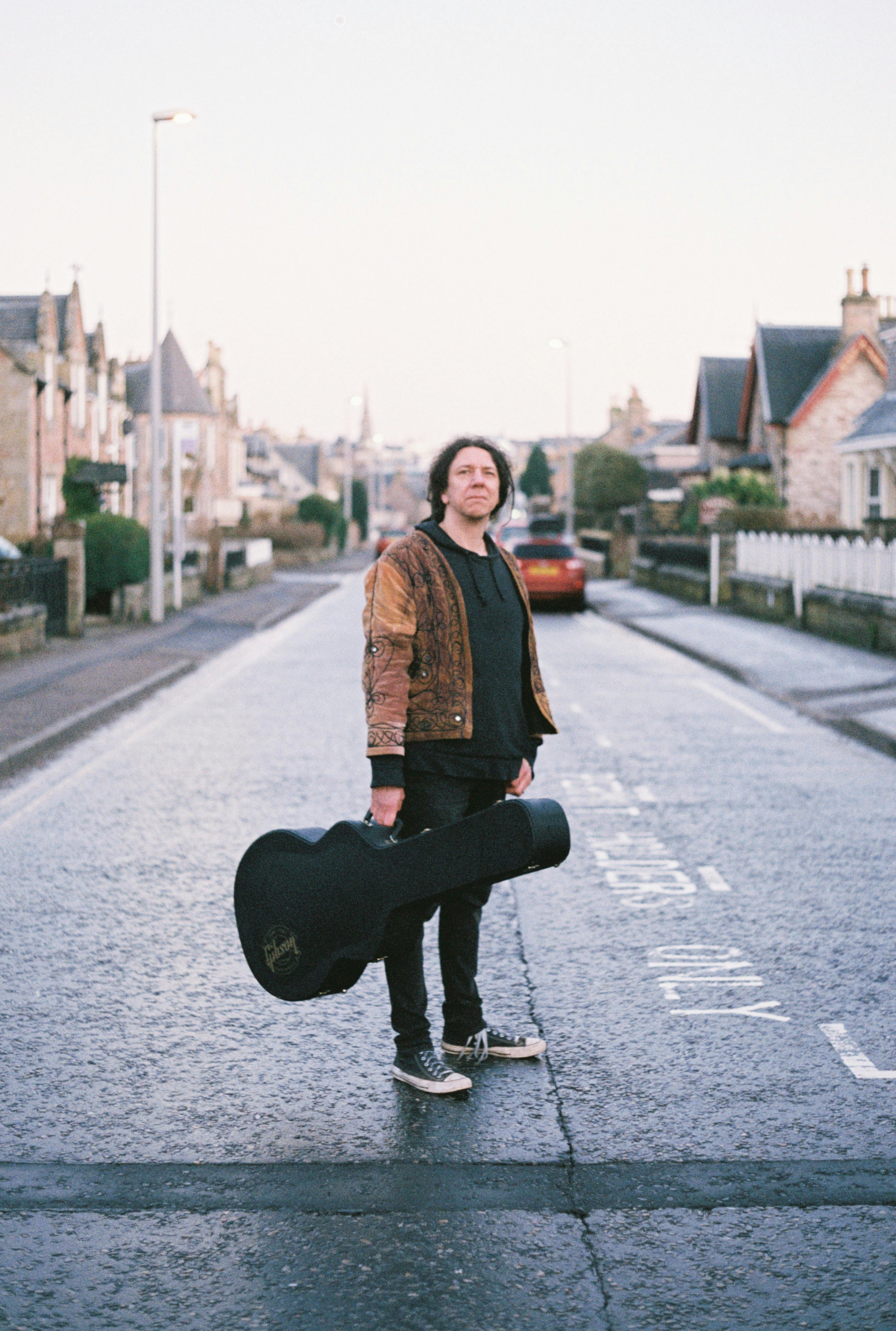 A musician stands confidently on an Inverness street holding a guitar case.