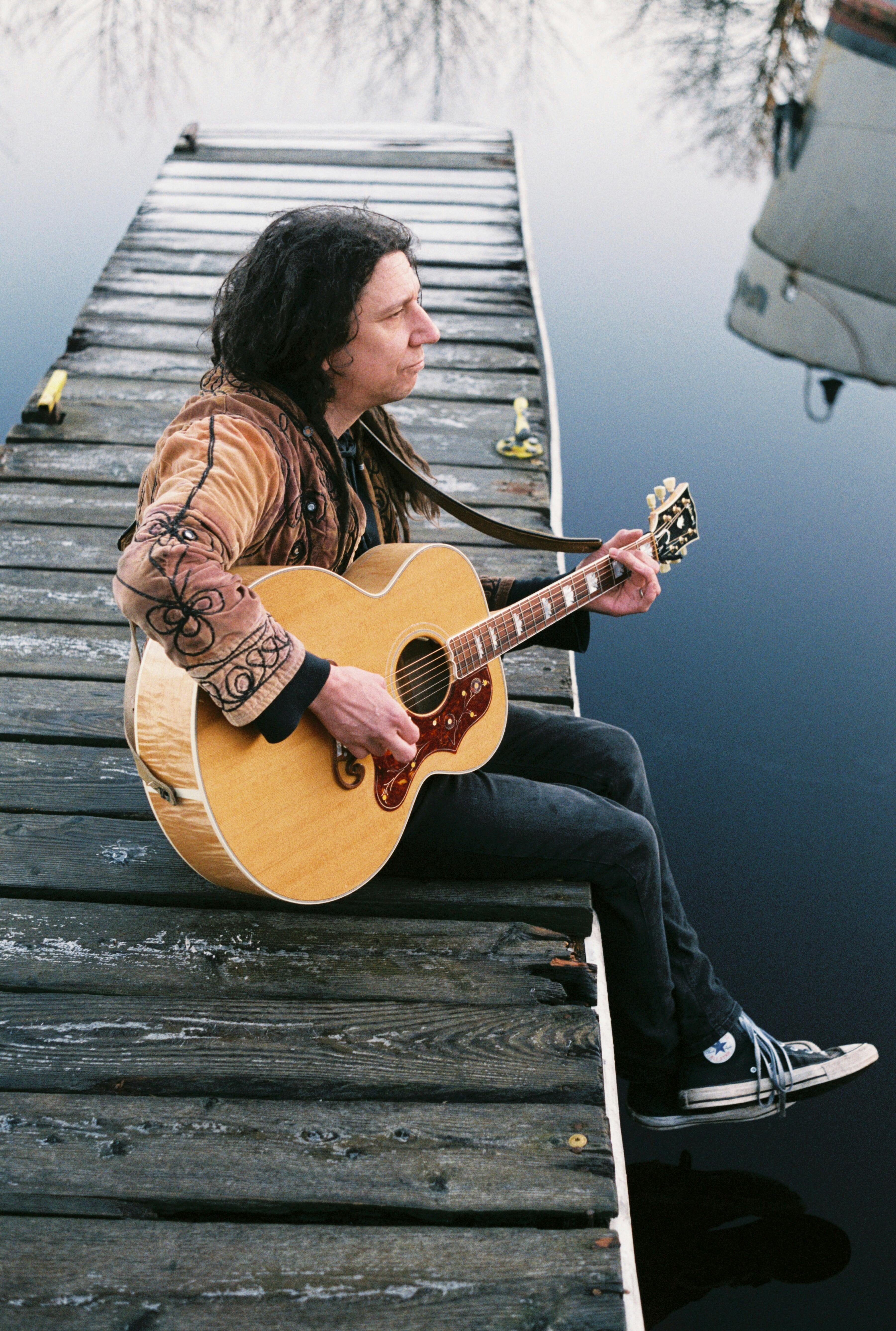 Musician playing acoustic guitar while sitting on a wooden dock in Inverness, Scotland.