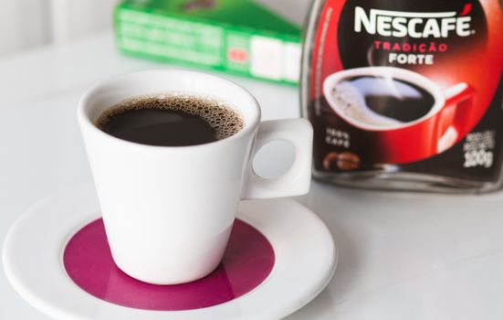 A modern white mug with black coffee sits on a pink saucer, Nescafé jar in the background.