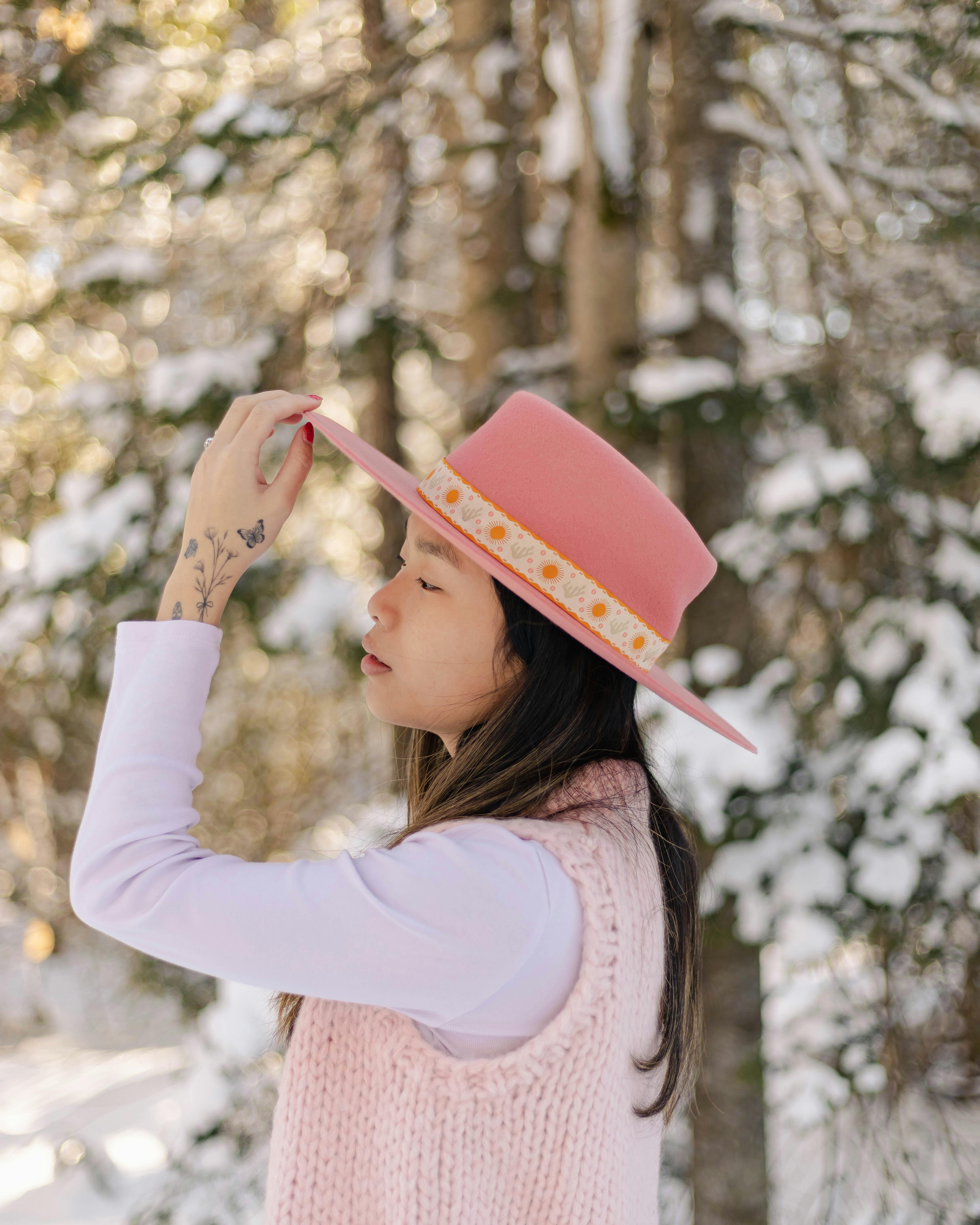 A woman in a pink hat touches the brim, surrounded by a snowy forest in winter.