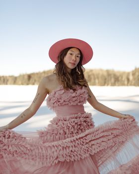 A woman in a pink dress and hat poses artistically against a snowy winter backdrop.