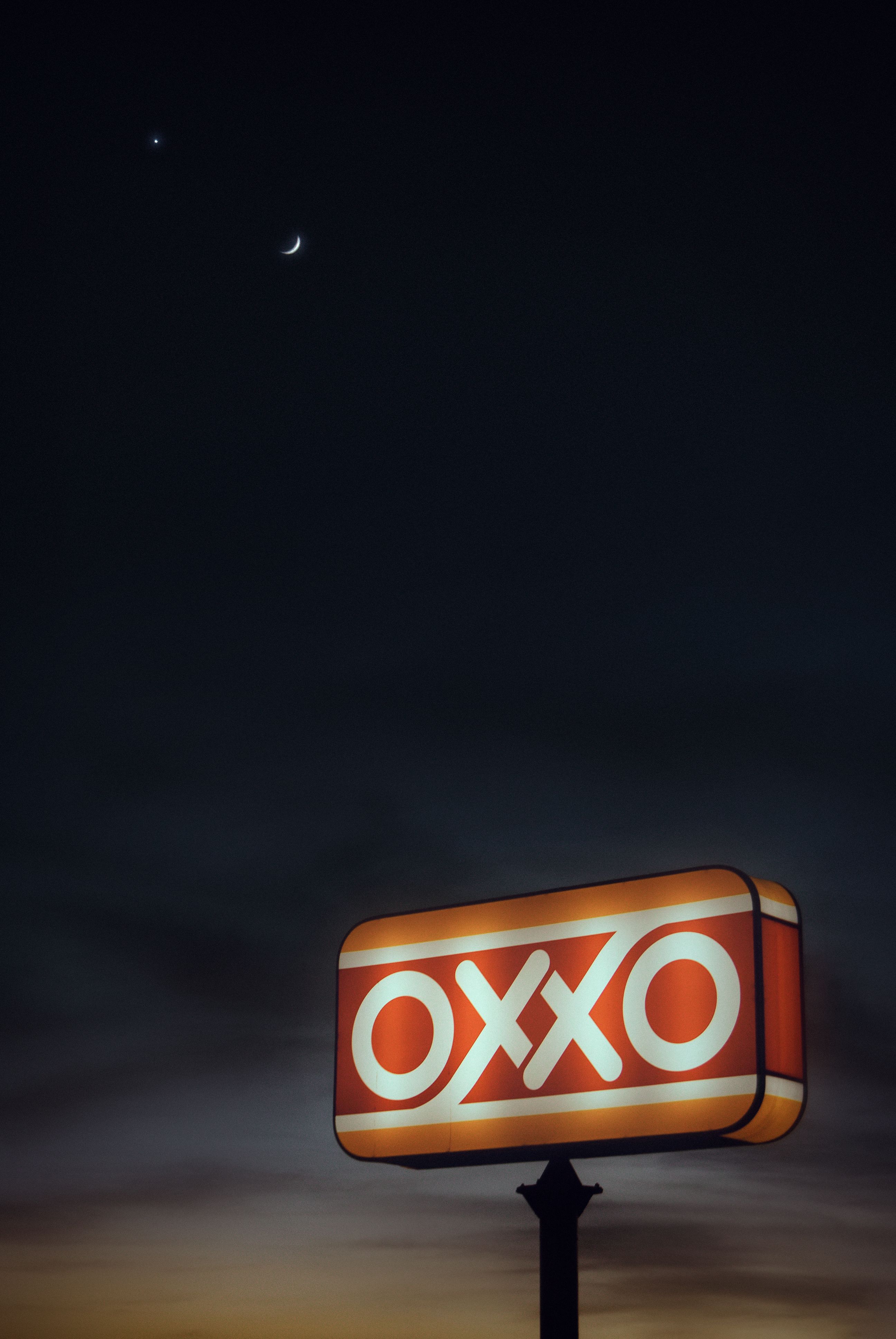 Illuminated OXXO sign with crescent moon and stars in the dark evening sky.