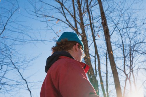 Free A man in a red jacket and blue beanie enjoys the outdoors on a crisp fall day, with bare trees against a clear sky. Stock Photo