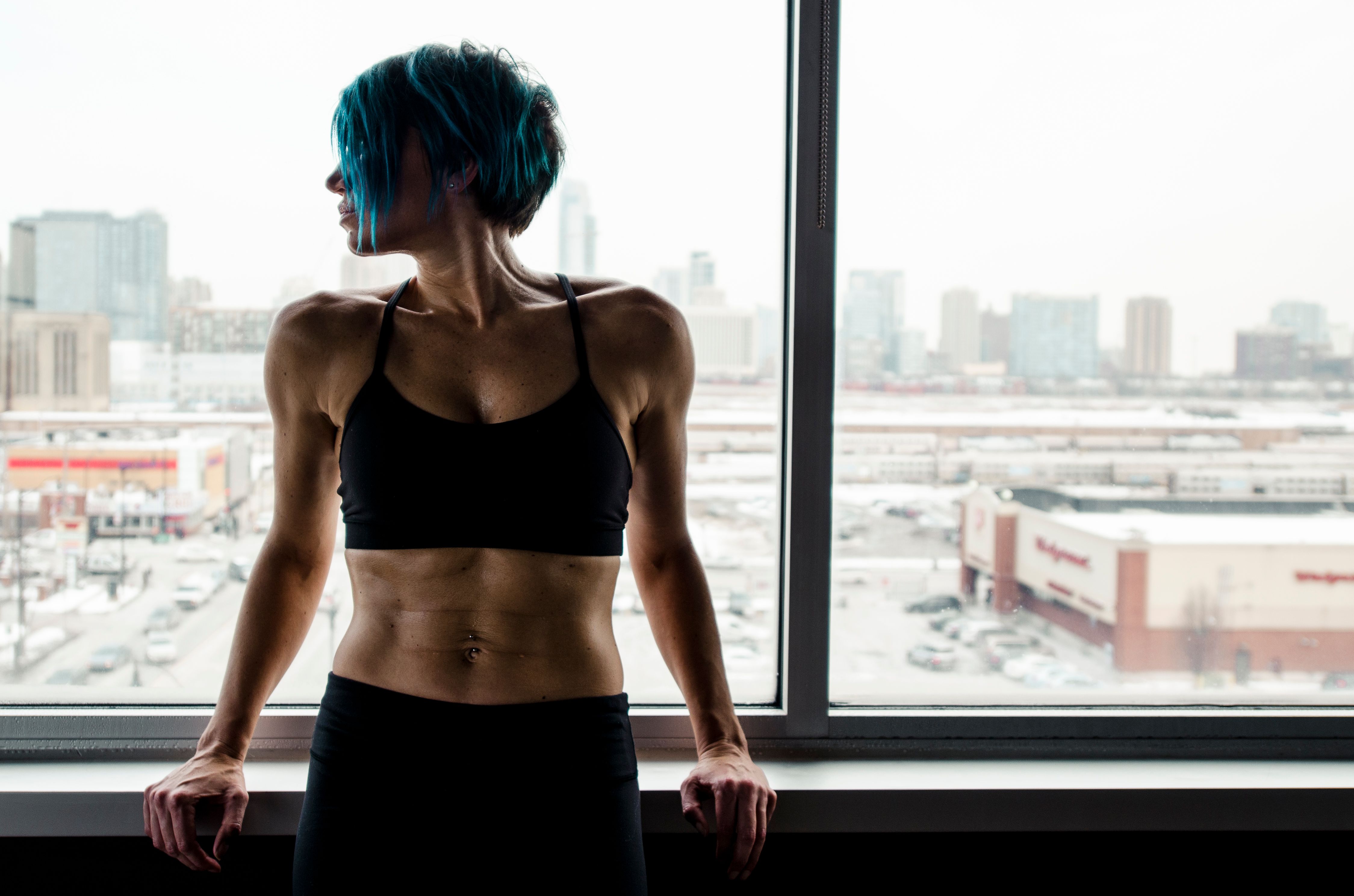 Fit woman with blue hair leans against a window overlooking an urban landscape.