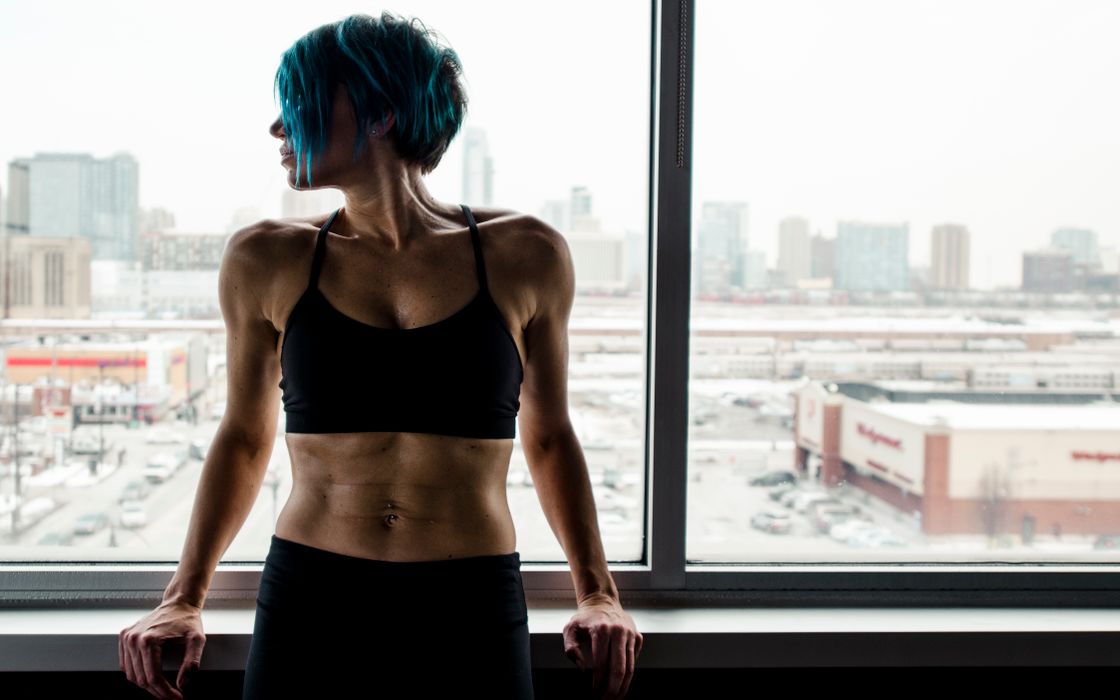 Fit woman with blue hair leans against a window overlooking an urban landscape.