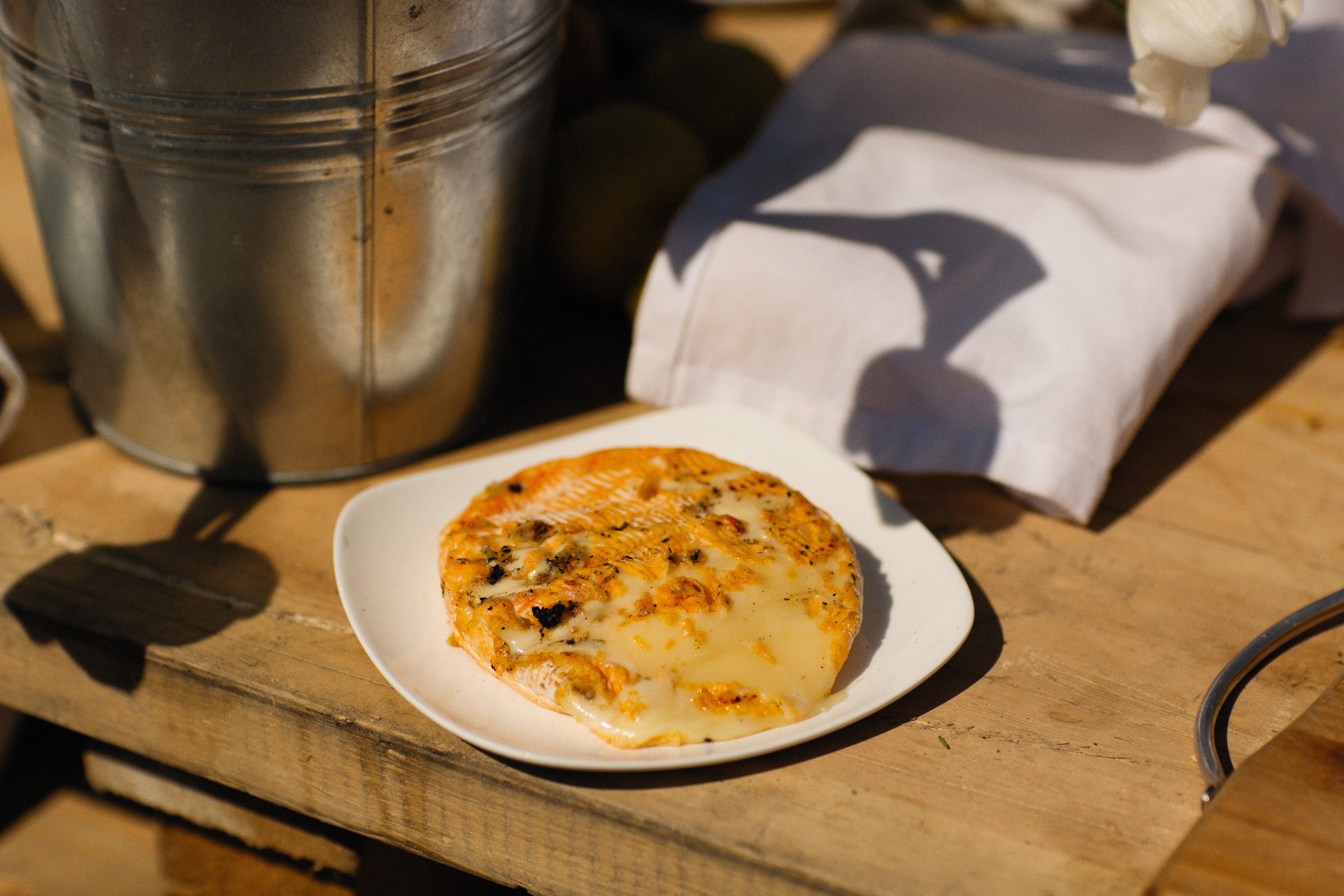 Galette de pain gratinée au fromage fondu sur une assiette blanche, posée sur une planche de bois en plein soleil