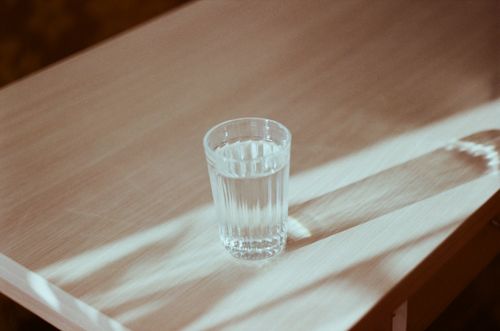 Free A glass of water casting a shadow on a wooden table illuminated by soft light. Stock Photo