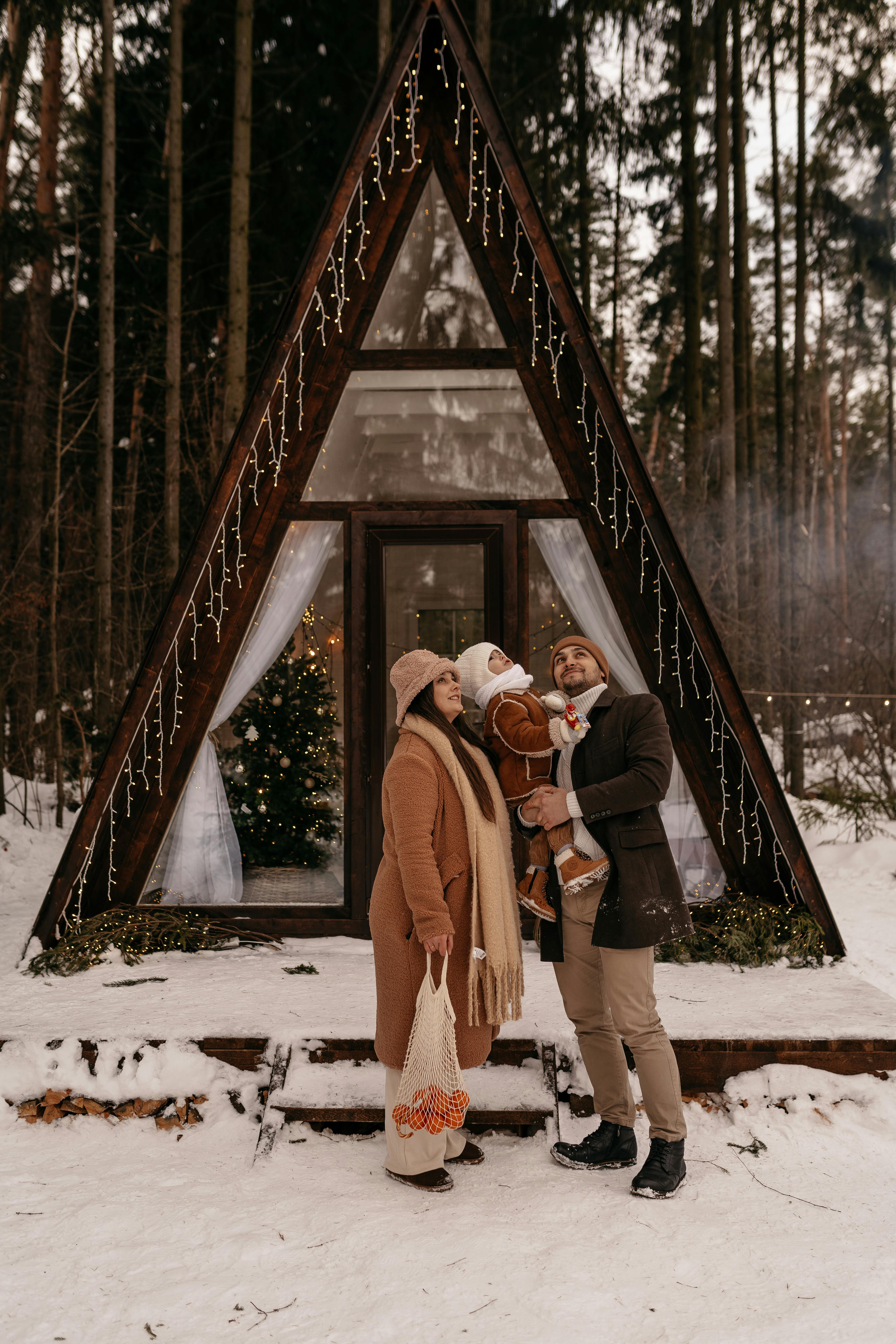 Couple with child outside a snowy A-frame cabin in a winter forest setting.