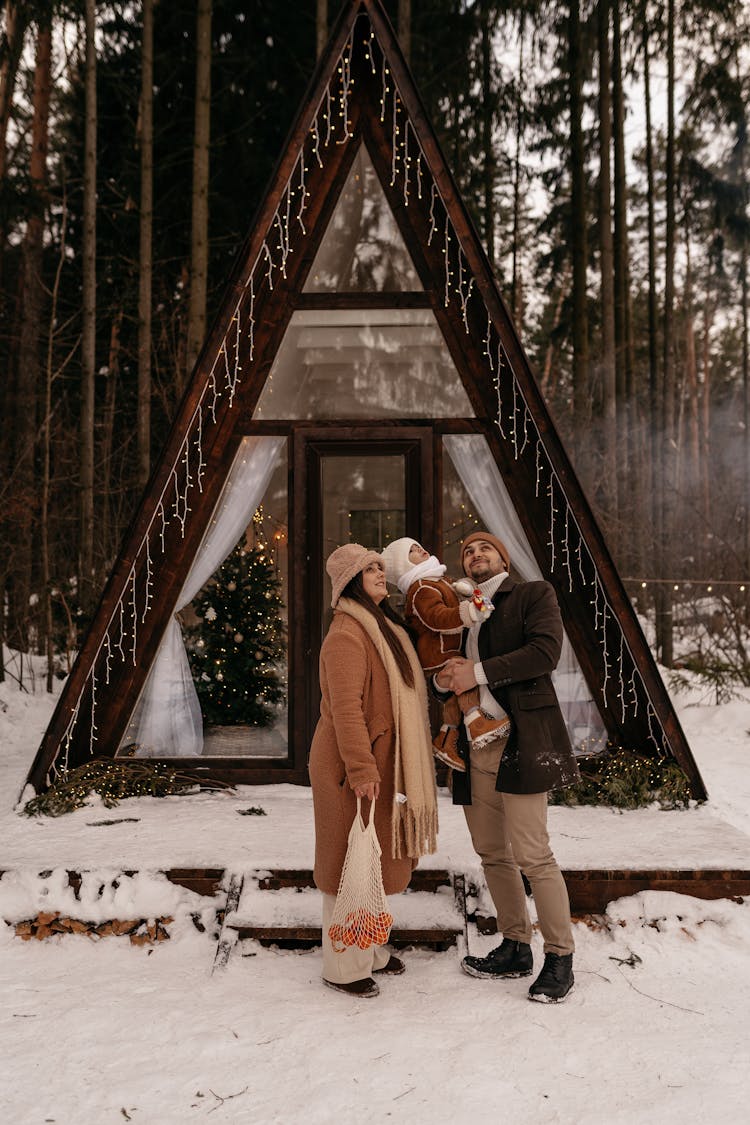 Couple With Child Standing In Snow In Front Of A-Frame Cabim