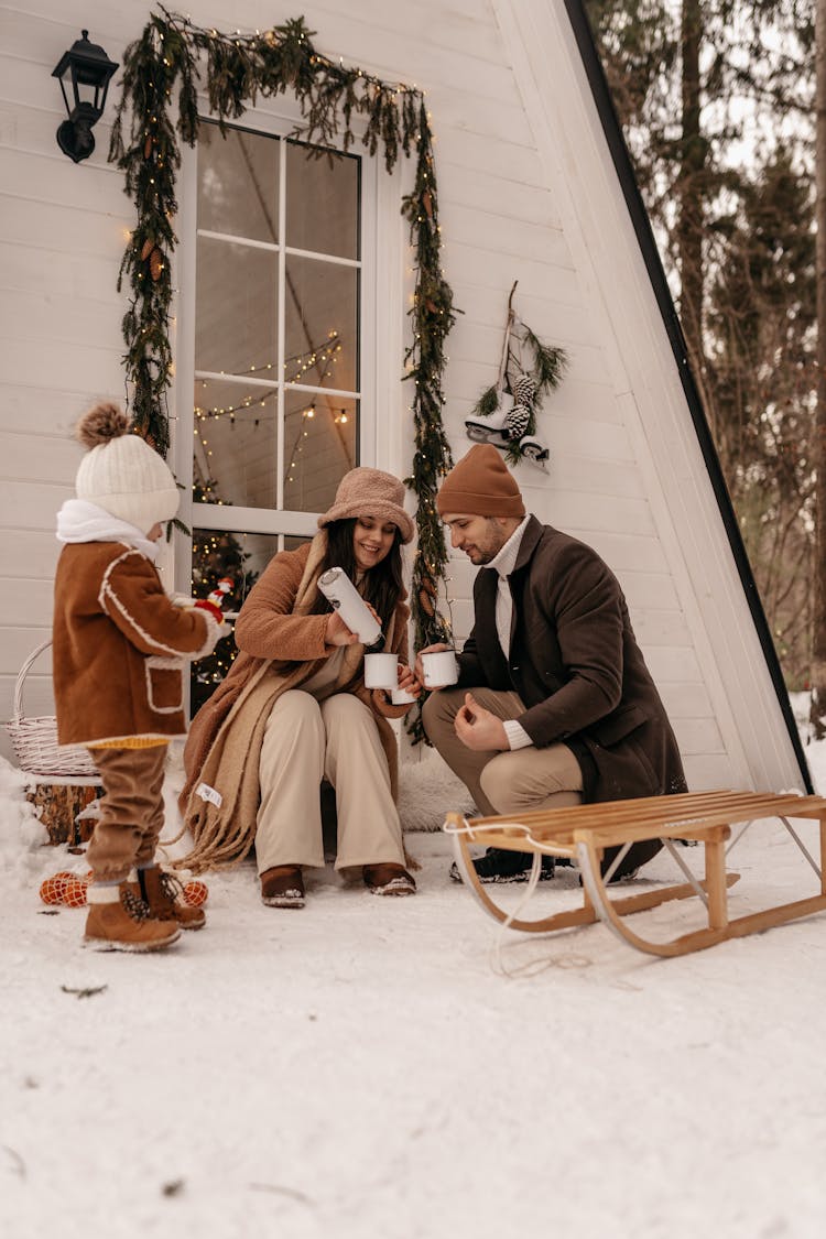 Couple With Child Sitting On Snow Drinking Tea From Mugs