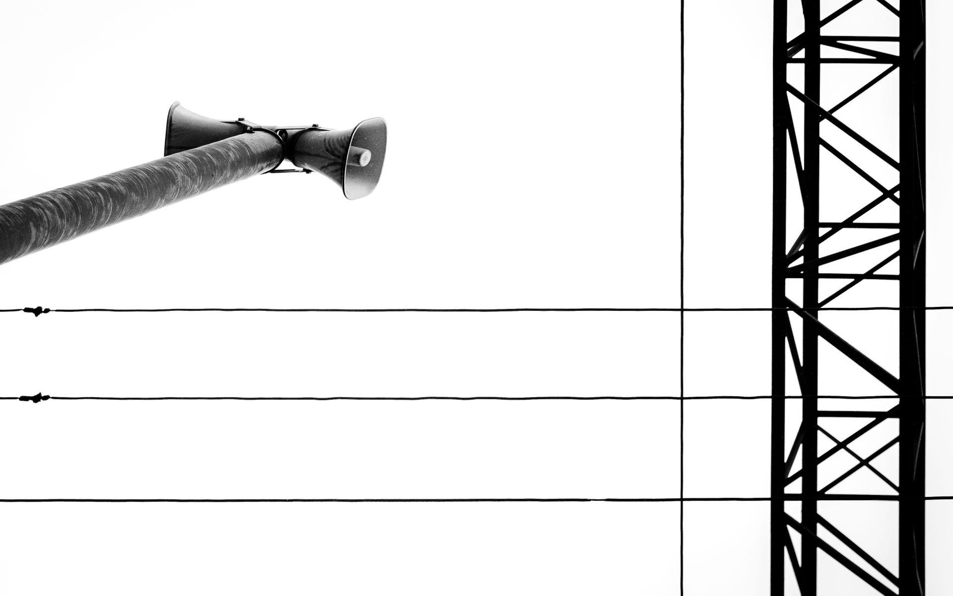 Monochrome image of a loudspeaker on a pole with power lines and a transmission tower.