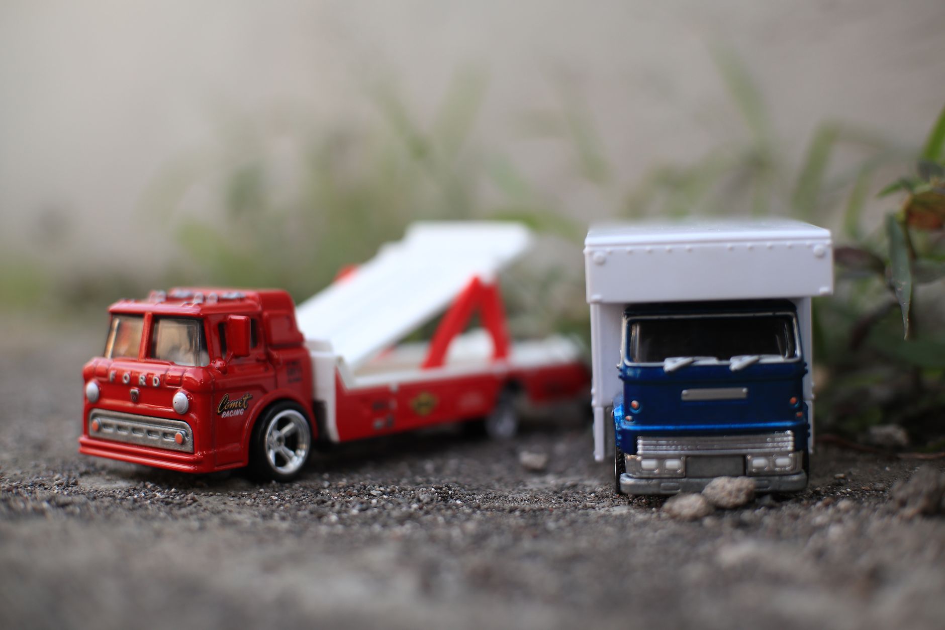 Two miniature toy trucks, one red and one blue, on a gravel surface outdoors.