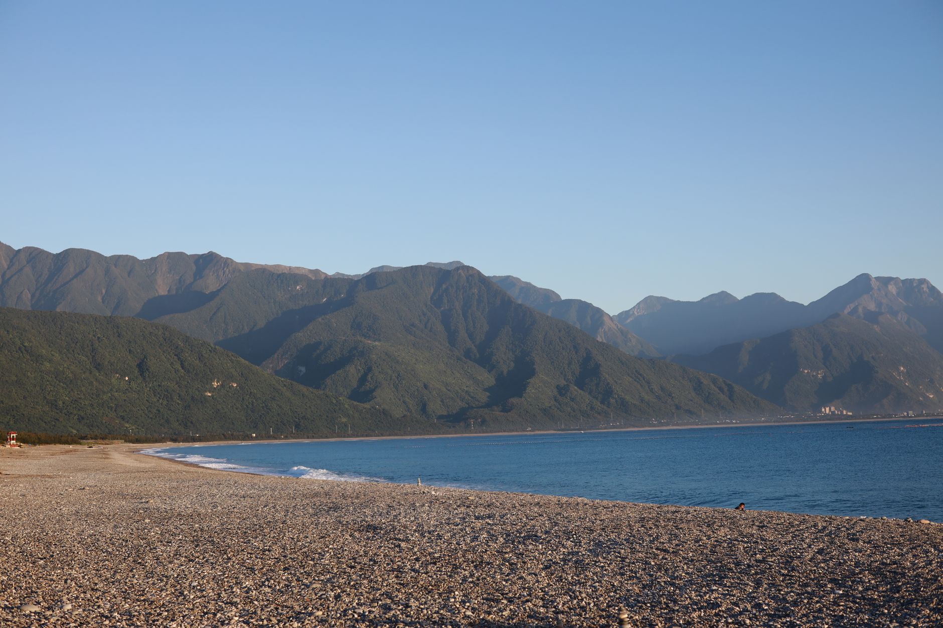 A tranquil pebble beach in Taiwan, framed by mountains and a clear blue sky.