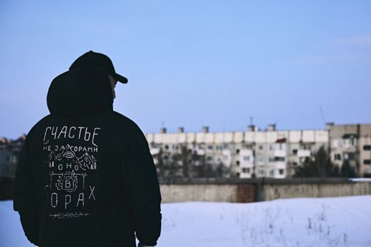 A man wearing a hoodie stands in front of snow-covered apartment buildings.