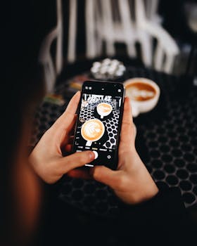 Hands photographing latte art with a mobile phone at a cafe.