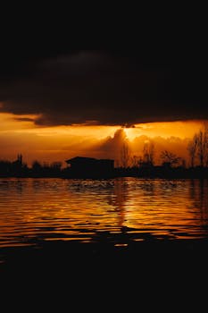 Silhouette of trees and dark clouds against a vibrant sunset reflecting on a calm lake.