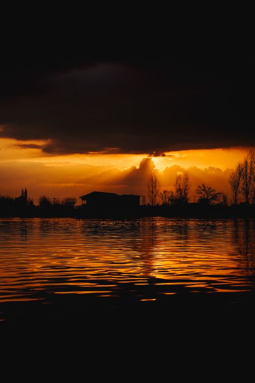 Free Silhouette of trees and dark clouds against a vibrant sunset reflecting on a calm lake. Stock Photo
