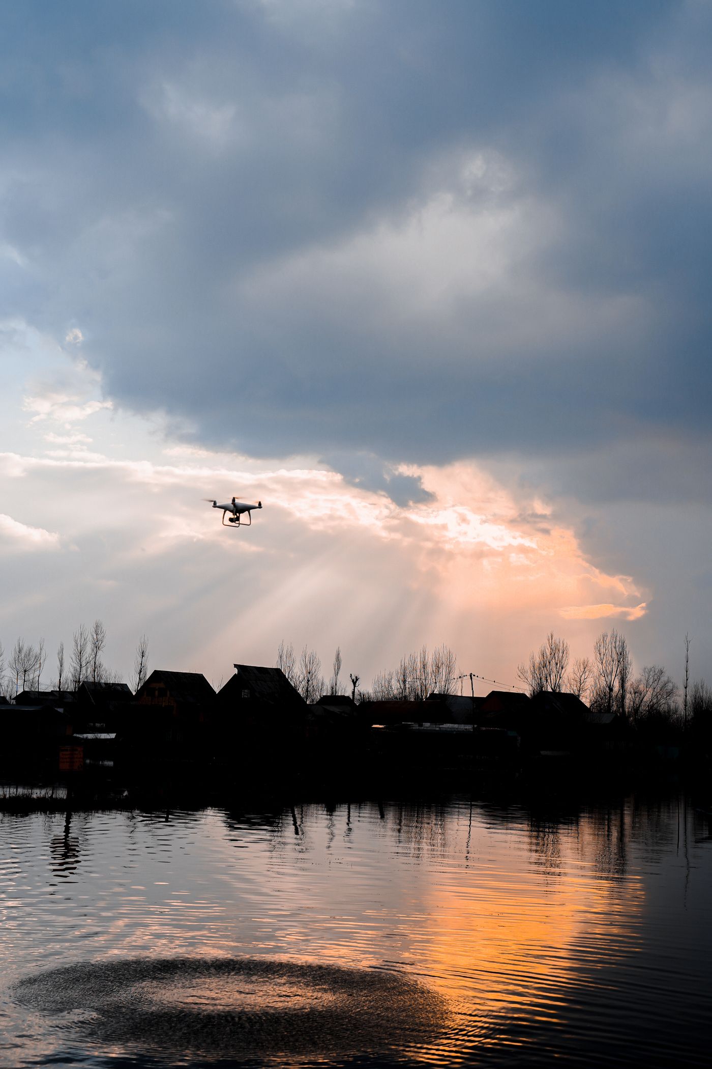 A small quadcopter drone hovering high above rooftops and bare trees, silhouetted against a bright break in heavy cloud at dusk