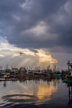 Drone flying over calm water near houses with dramatic clouds and sunlight breaking through.
