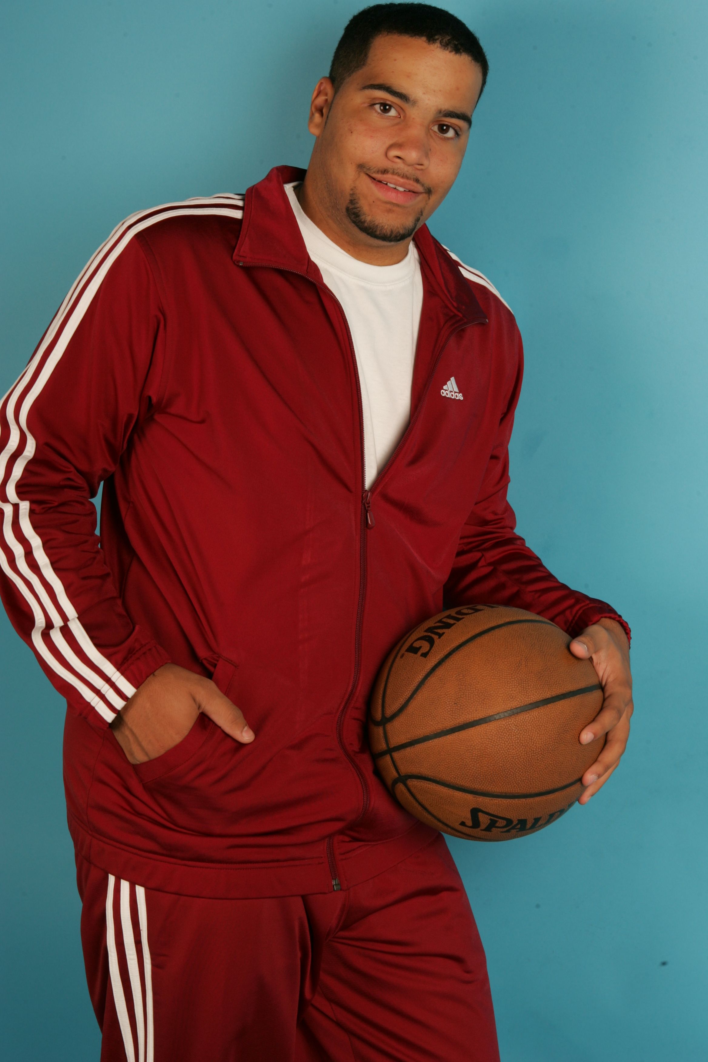 Adult male in a red tracksuit holding a basketball against a blue background.