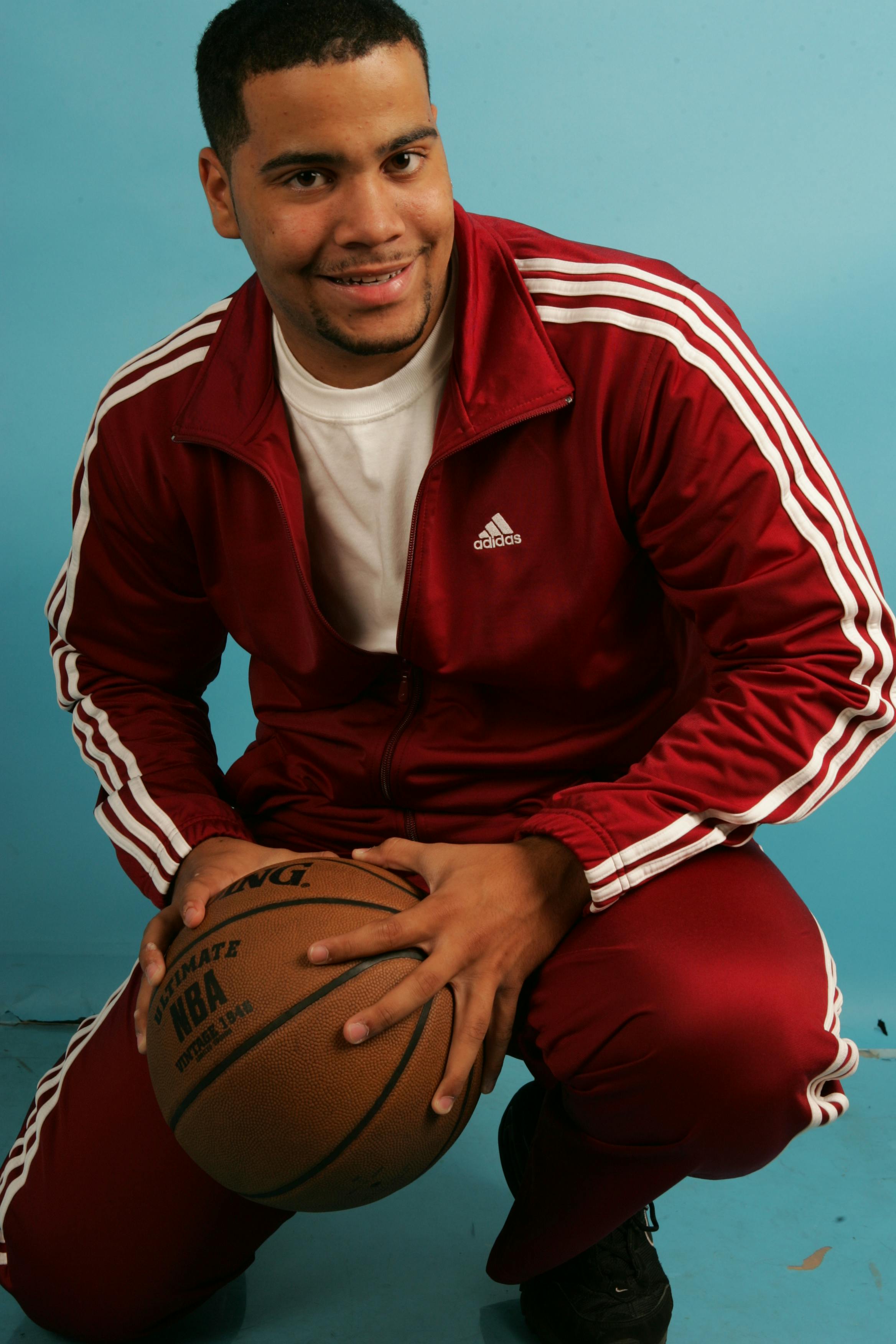 Smiling man in red Adidas tracksuit holding a basketball, set against a blue background indoors.