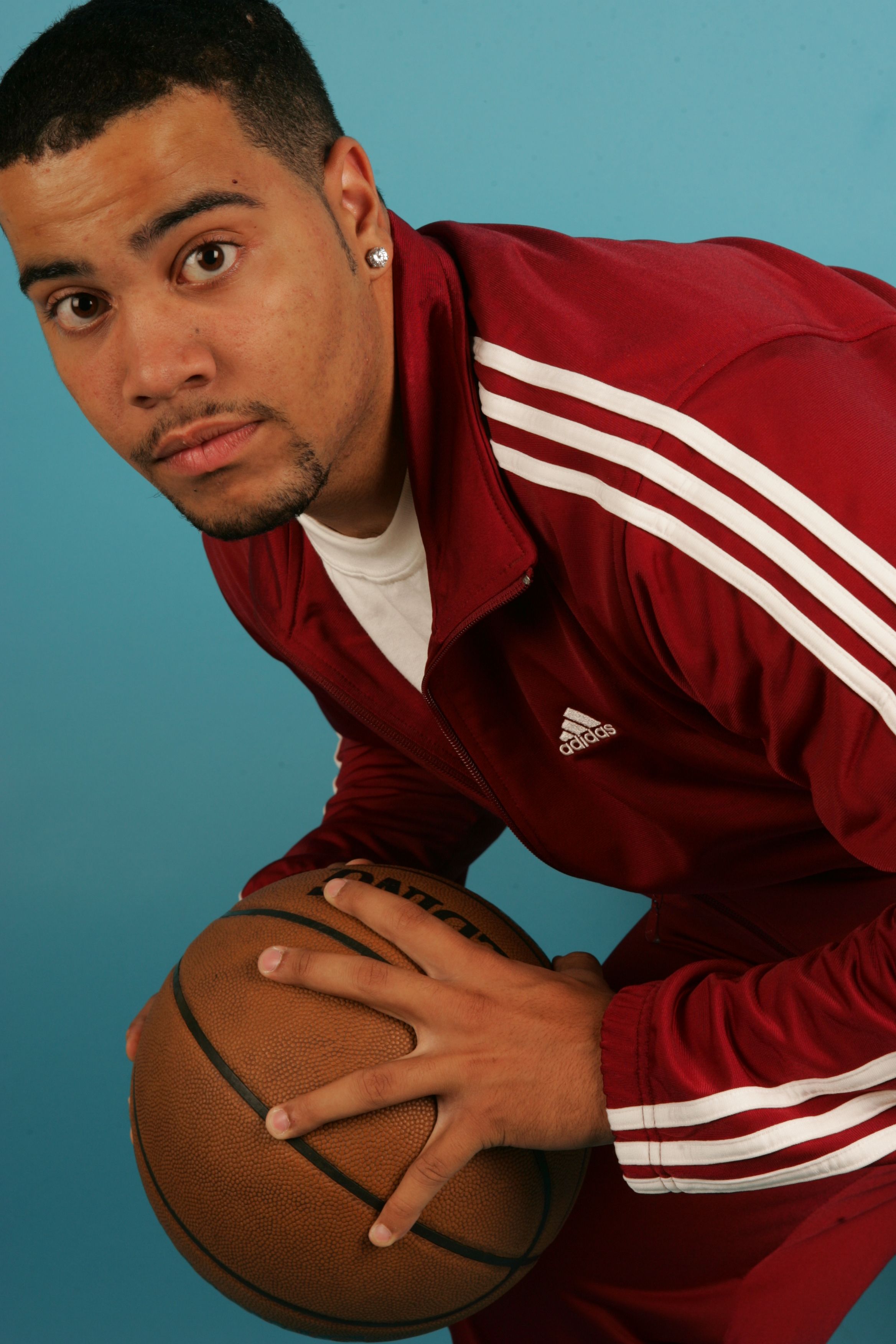 Focused young athlete holding a basketball in a red tracksuit against a blue background.