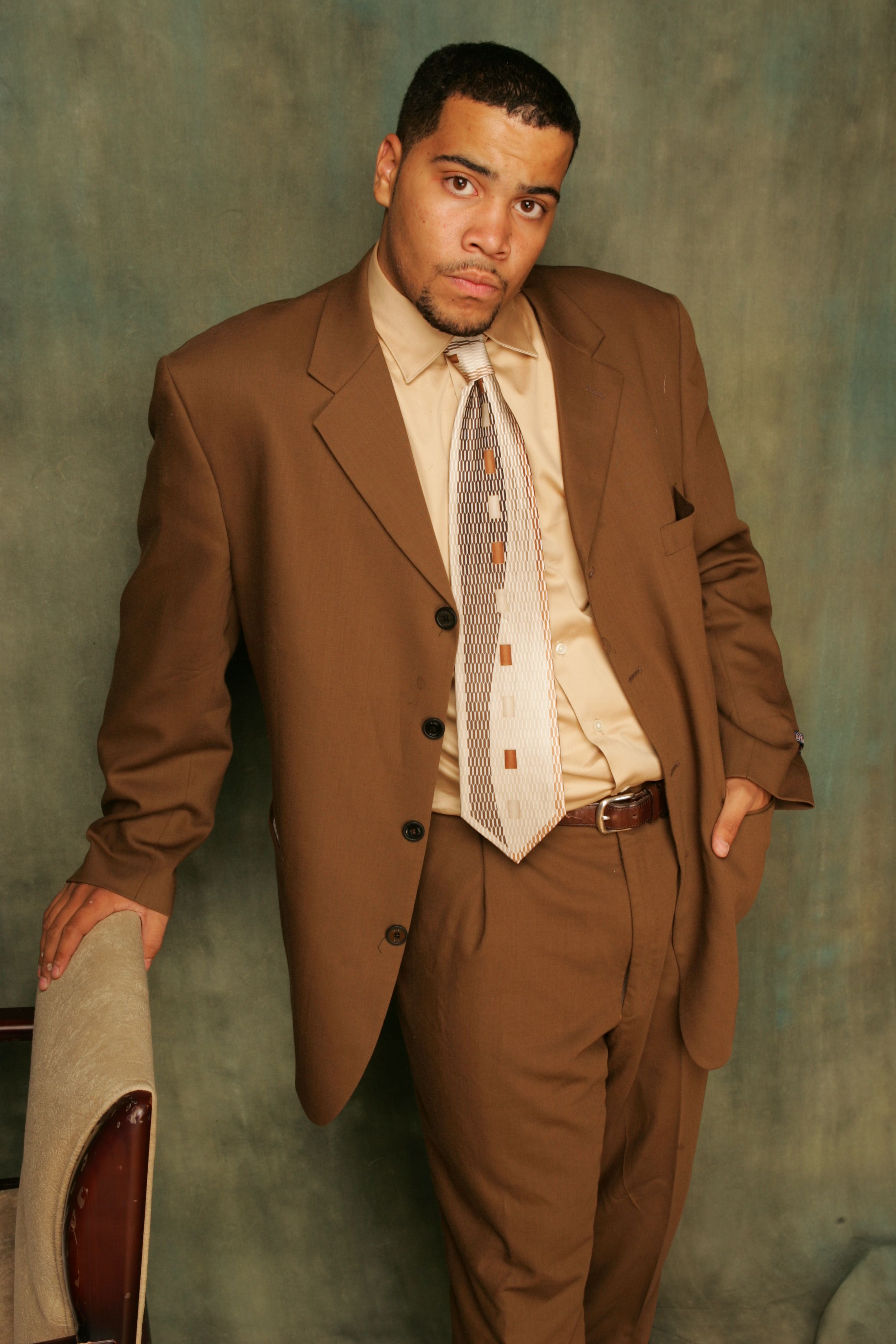 Portrait of a man in a brown suit standing confidently indoors with a hand in his pocket.