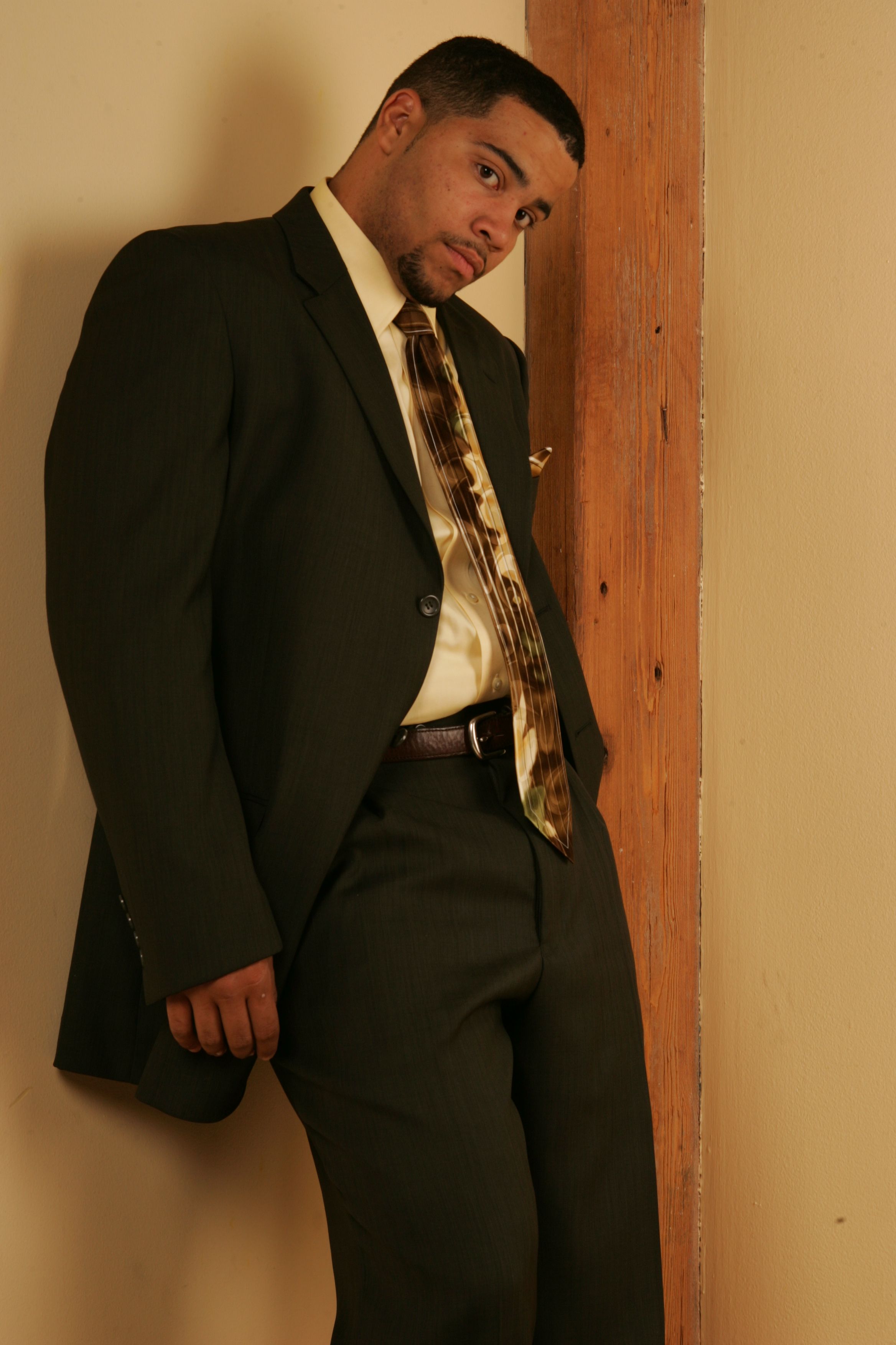 A confident man in a formal suit poses stylishly, leaning against a wall indoors.