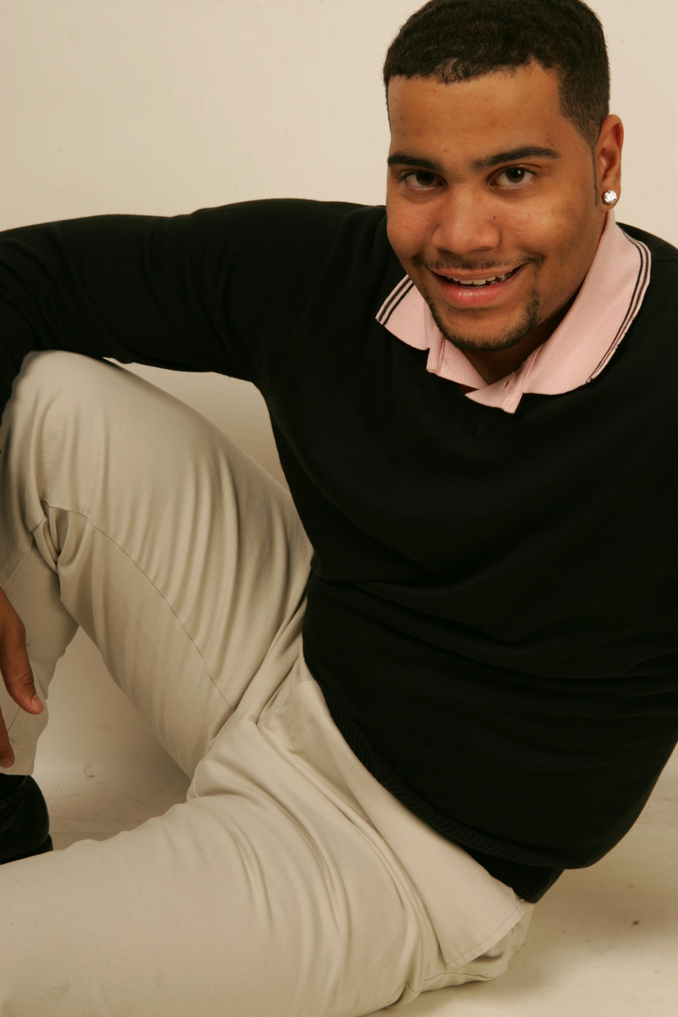 Portrait of a smiling man sitting indoors wearing a black sweater and white pants.