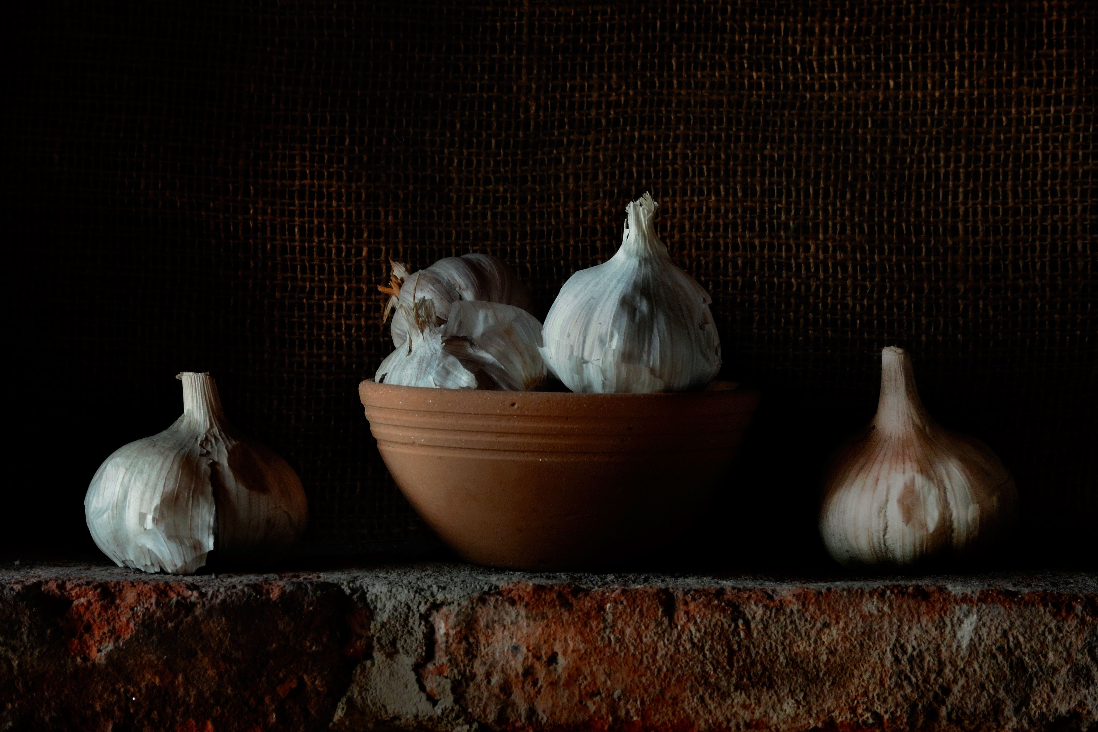 A moody still life photo featuring garlic bulbs in a clay bowl on a brick surface.