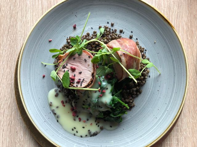 Elegant plated dish featuring bacon-wrapped pork medallions on a bed of lentils and greens.