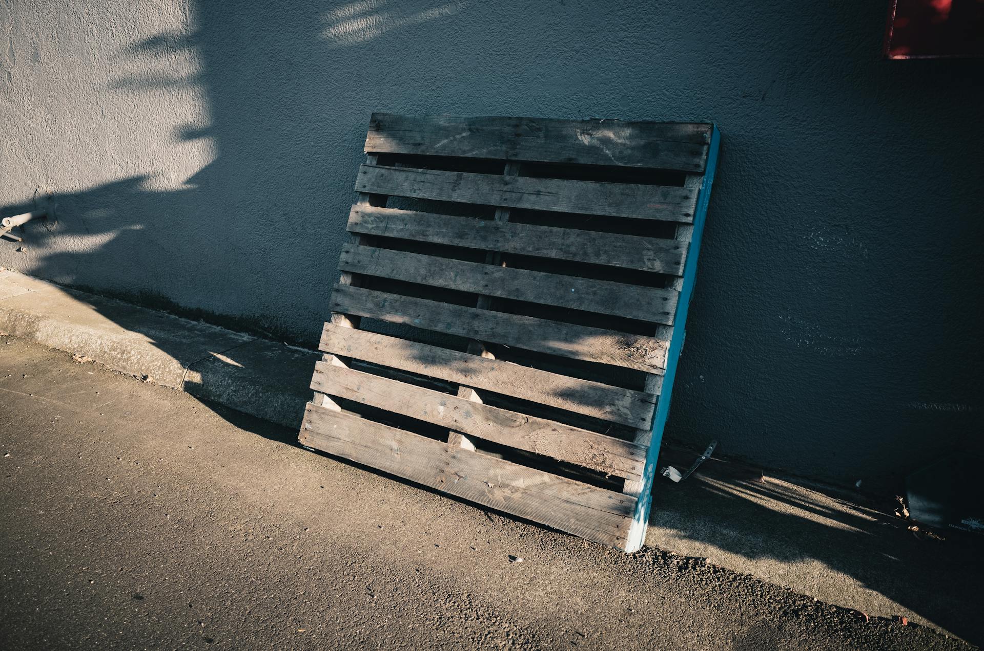 A wooden pallet leaning against a concrete wall casting shadows in daylight.